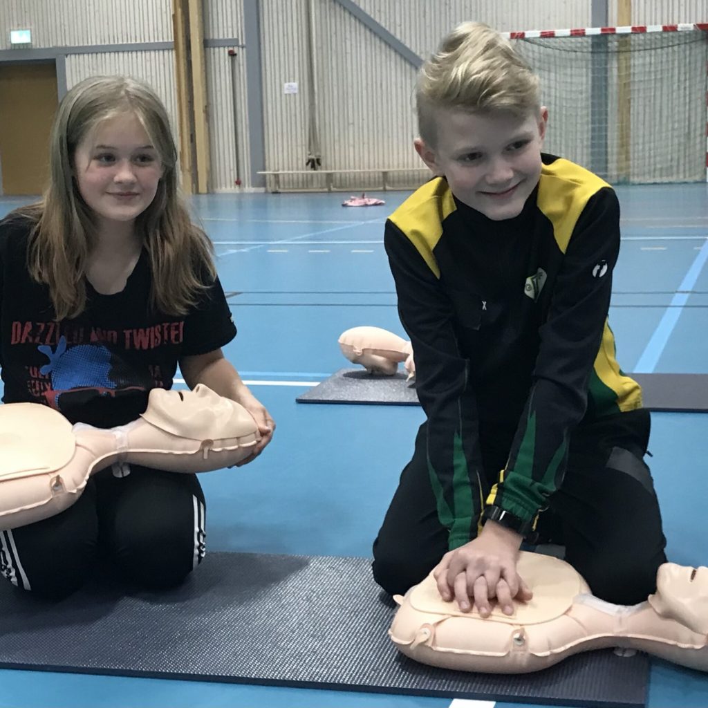 Två barn övar hjärt-lungräddning (HLR) på övningsdockor i en idrottshall